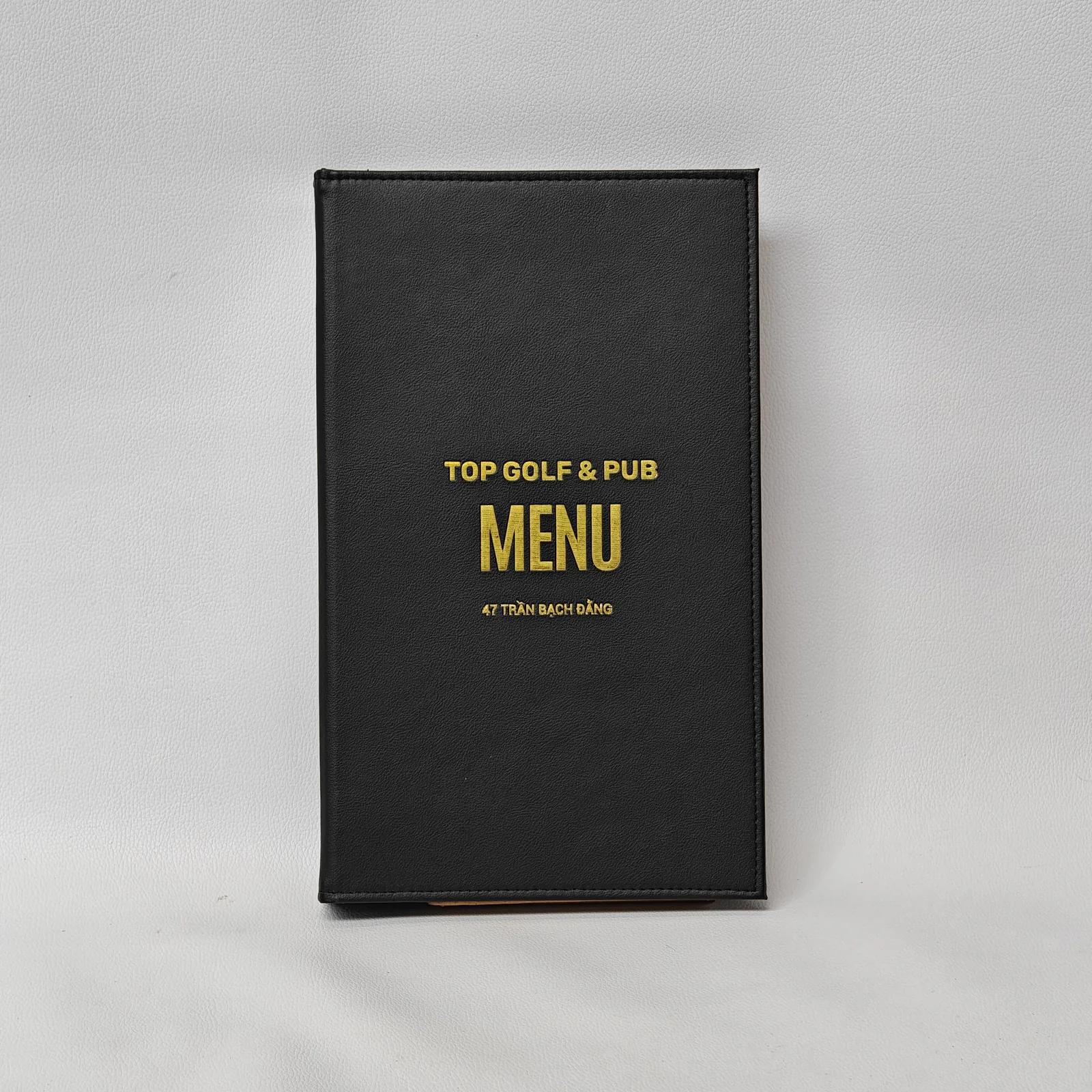Front view of a black textured leather menu cover with gold foil text and perimeter stitching.