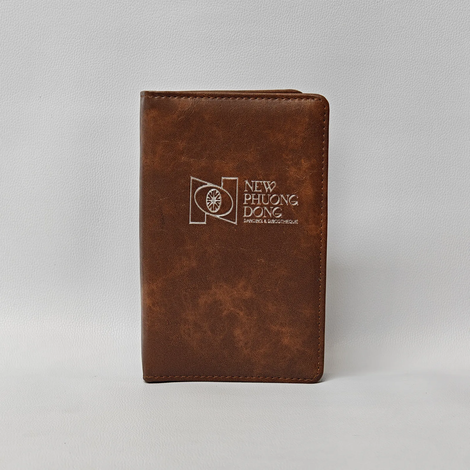 Front view of a closed brown textured leather menu cover with an embossed logo and perimeter stitching.