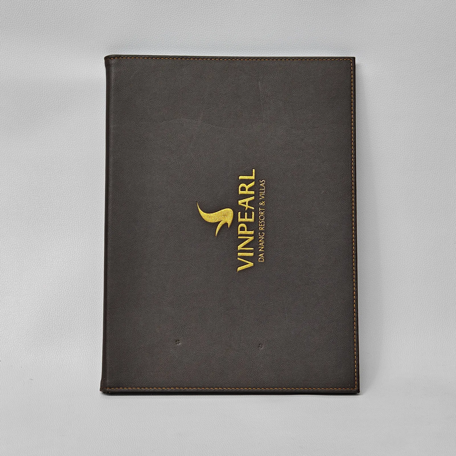 Front view of a closed dark brown leather folder with a gold logo and orange contrast stitching.