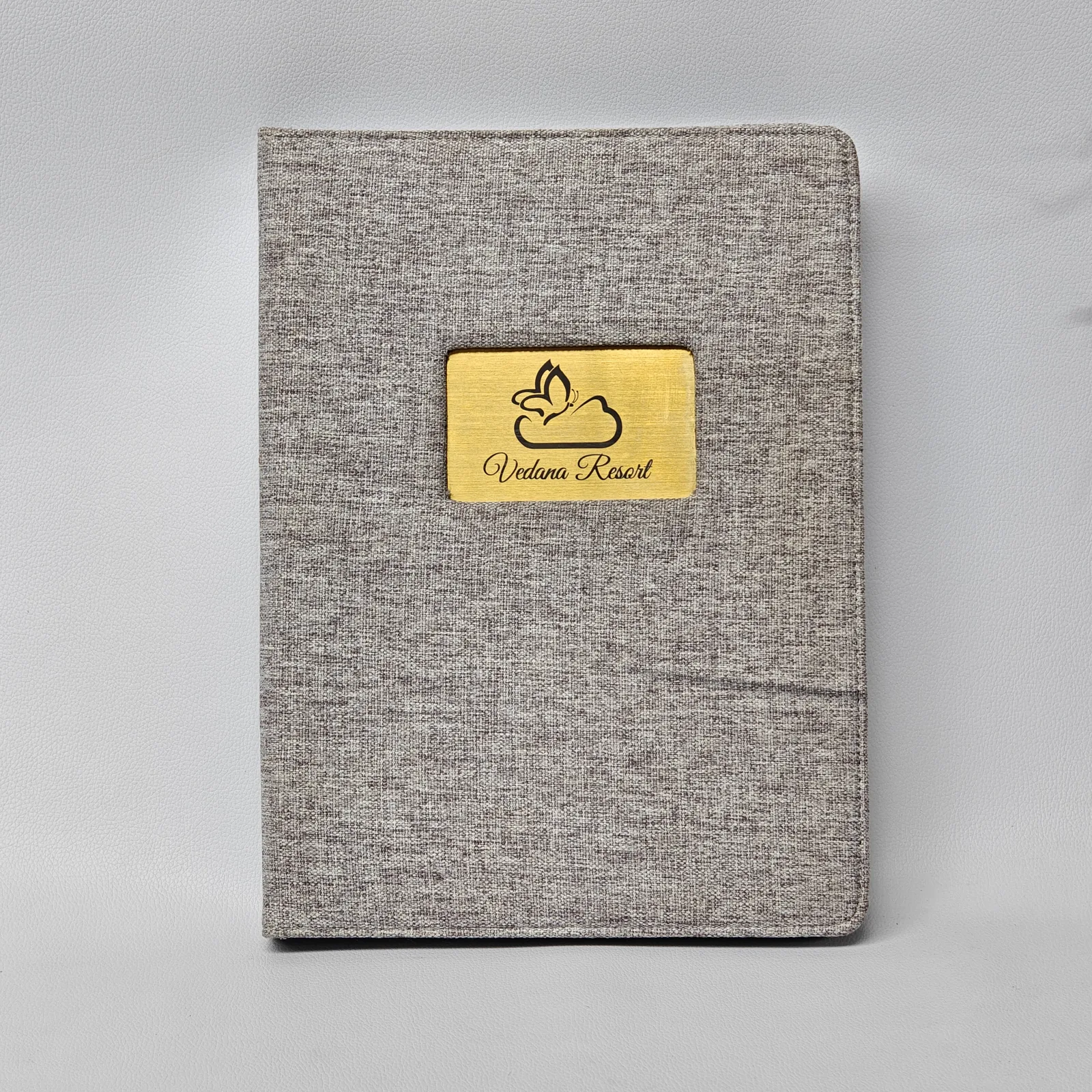Front view of a grey canvas menu cover featuring a central gold metal plate with an engraved logo.