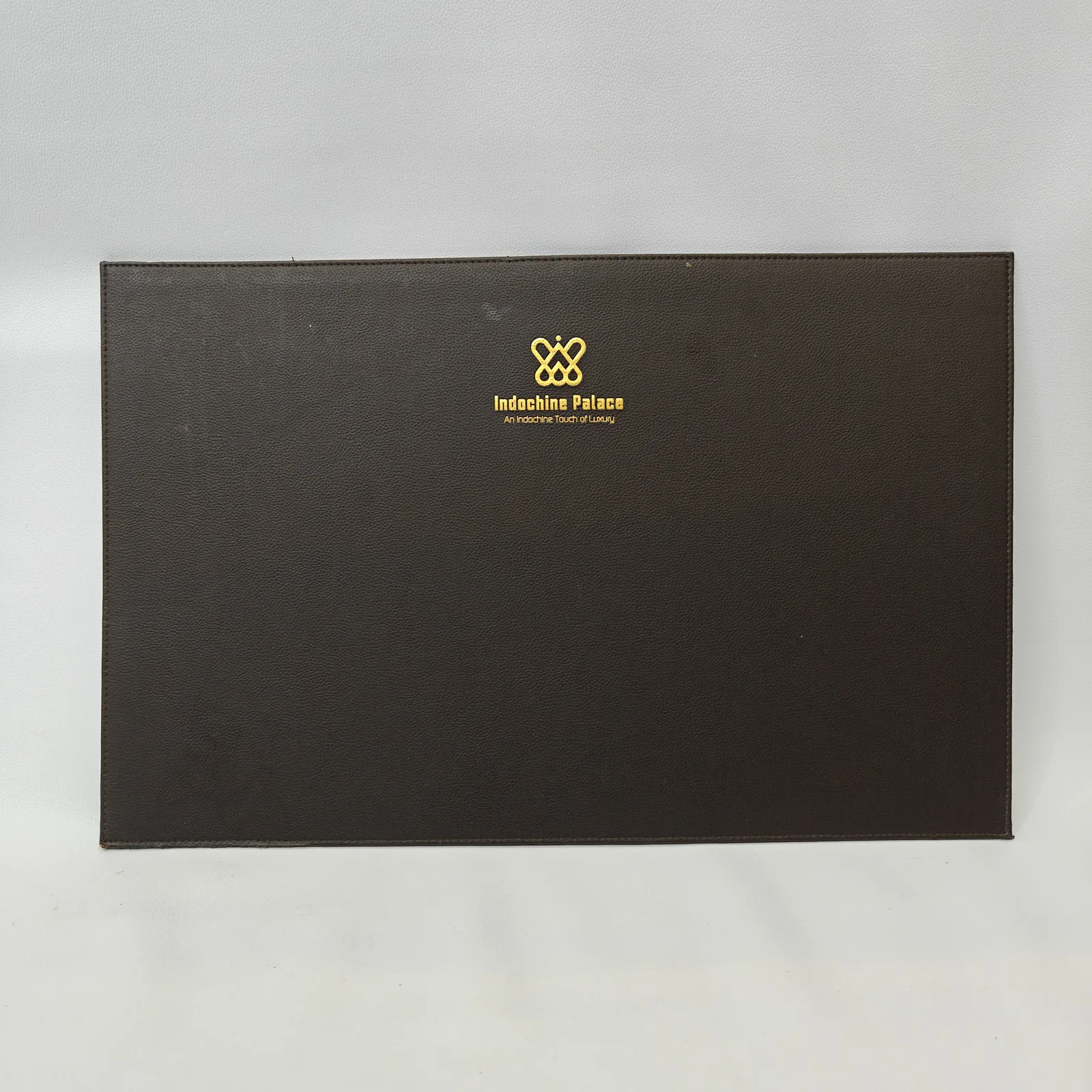 A rectangular dark brown leather desk mat with perimeter stitching and a gold logo in the center.