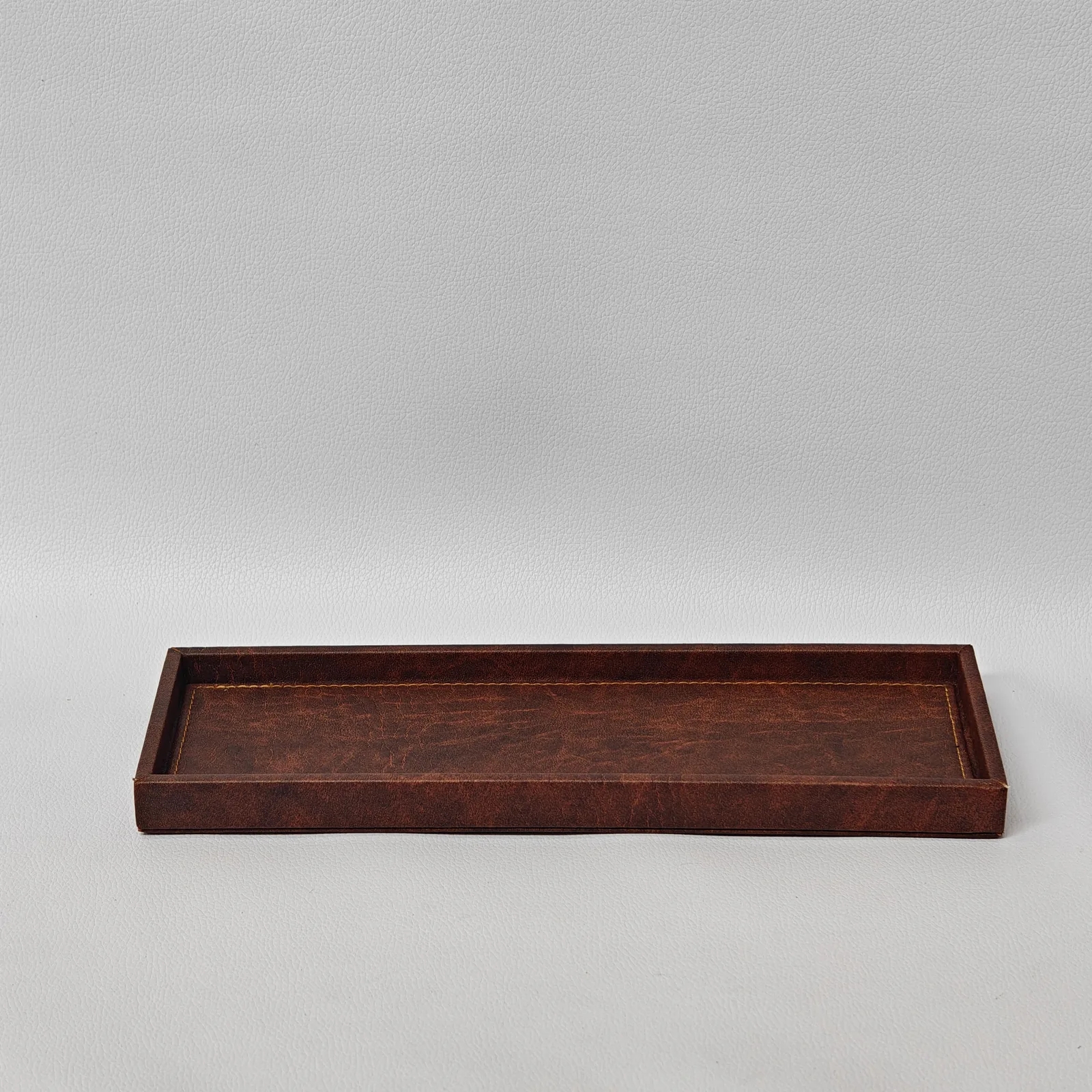 An angled view of a long, rectangular brown leather tray with raised edges and perimeter stitching.