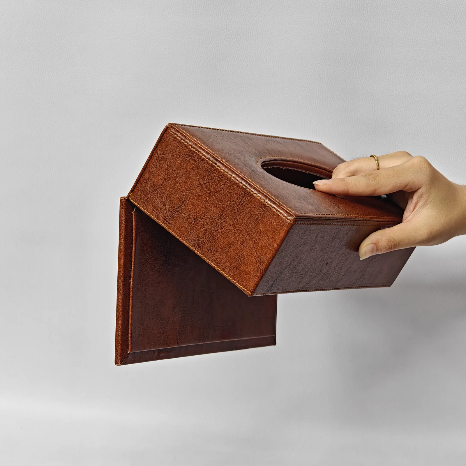 A hand opening the magnetic bottom flap of a textured brown leather tissue box cover.
