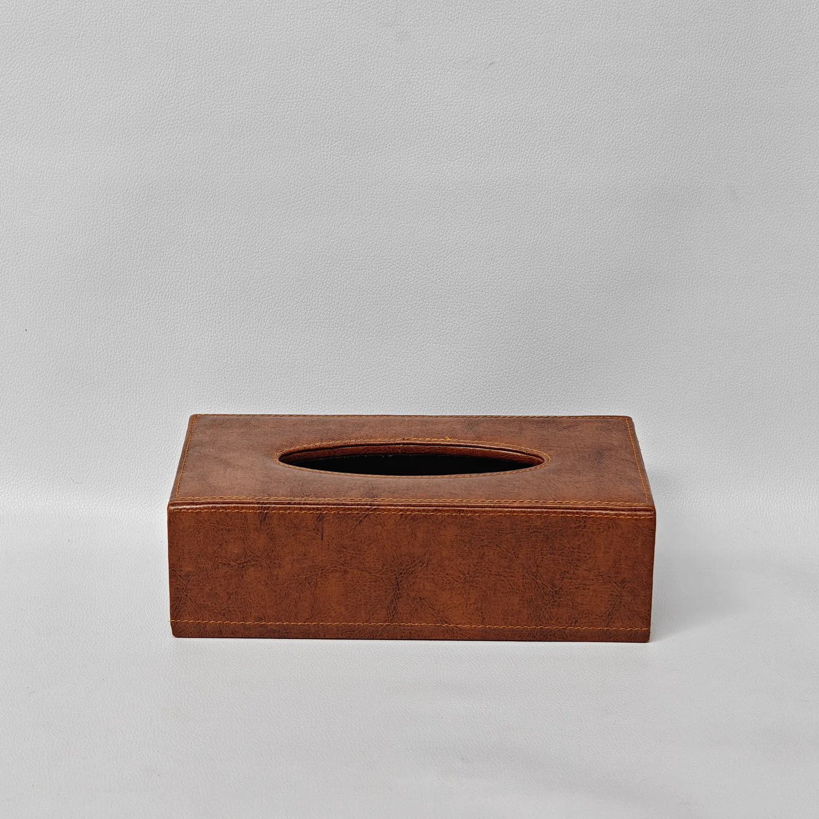 Front view of a textured brown leather tissue box cover with an oval top opening and perimeter stitching.