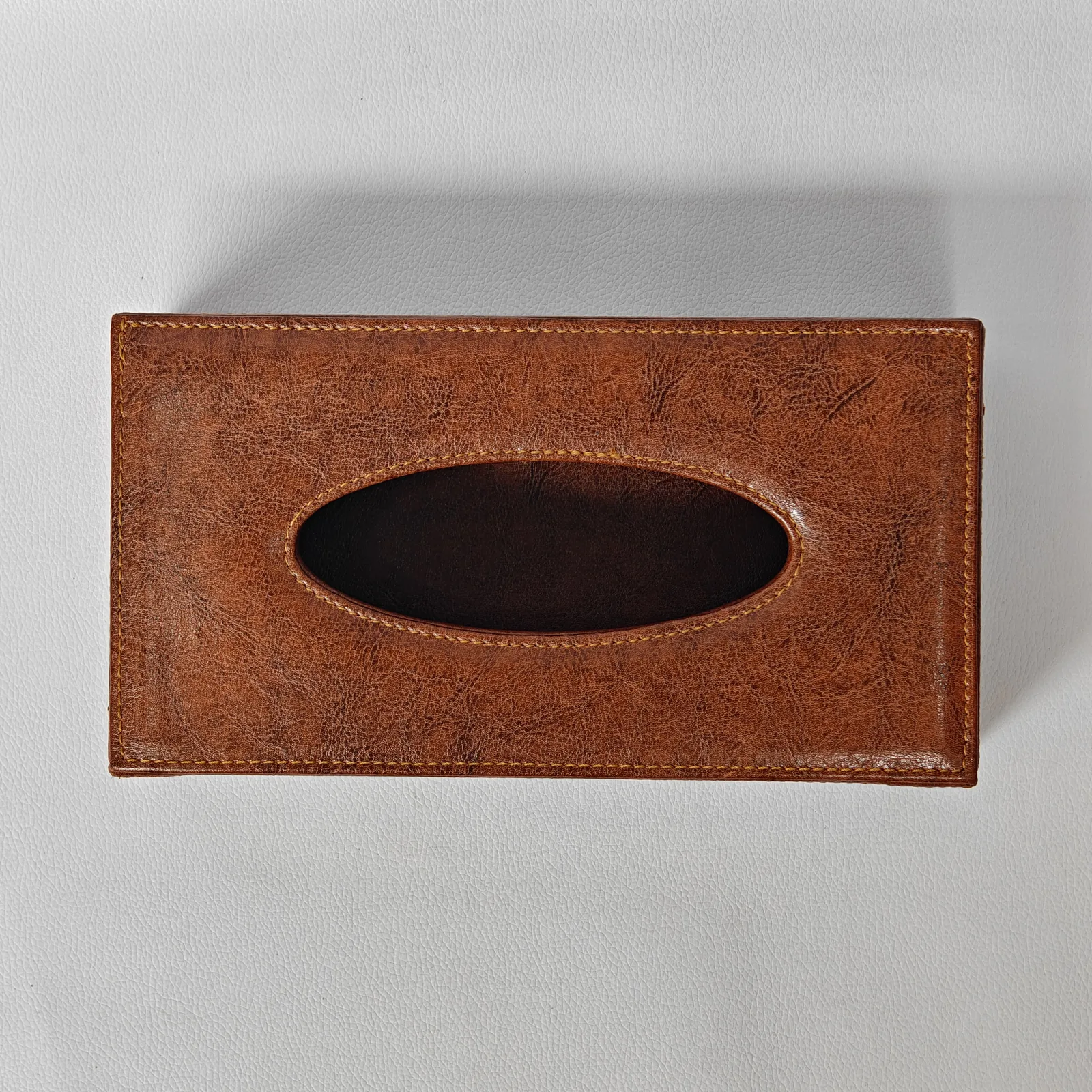 Top-down view of a textured brown leather tissue box cover showing the oval opening and decorative stitching.