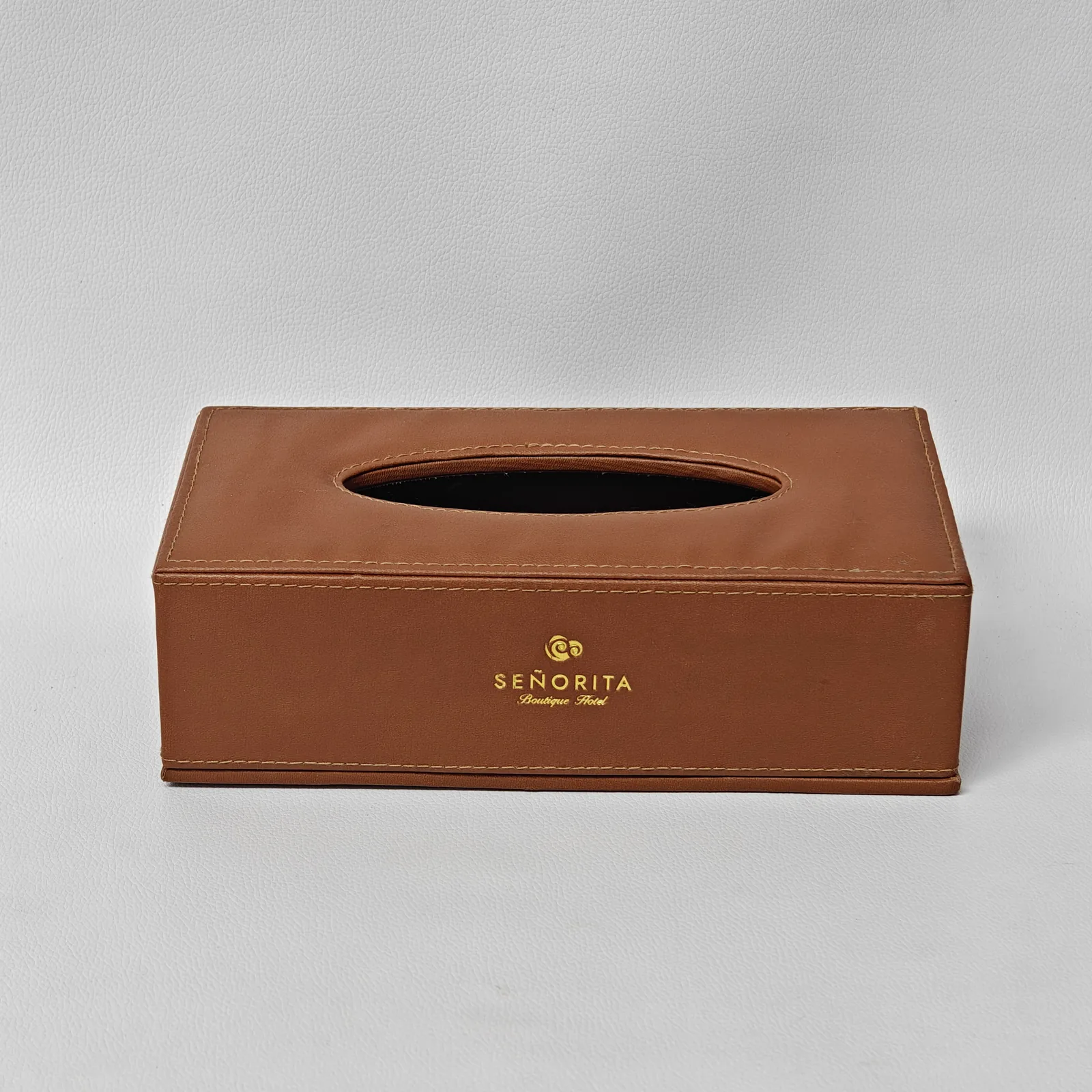 Front view of a smooth tan leather tissue box cover with a gold logo and an oval top opening.