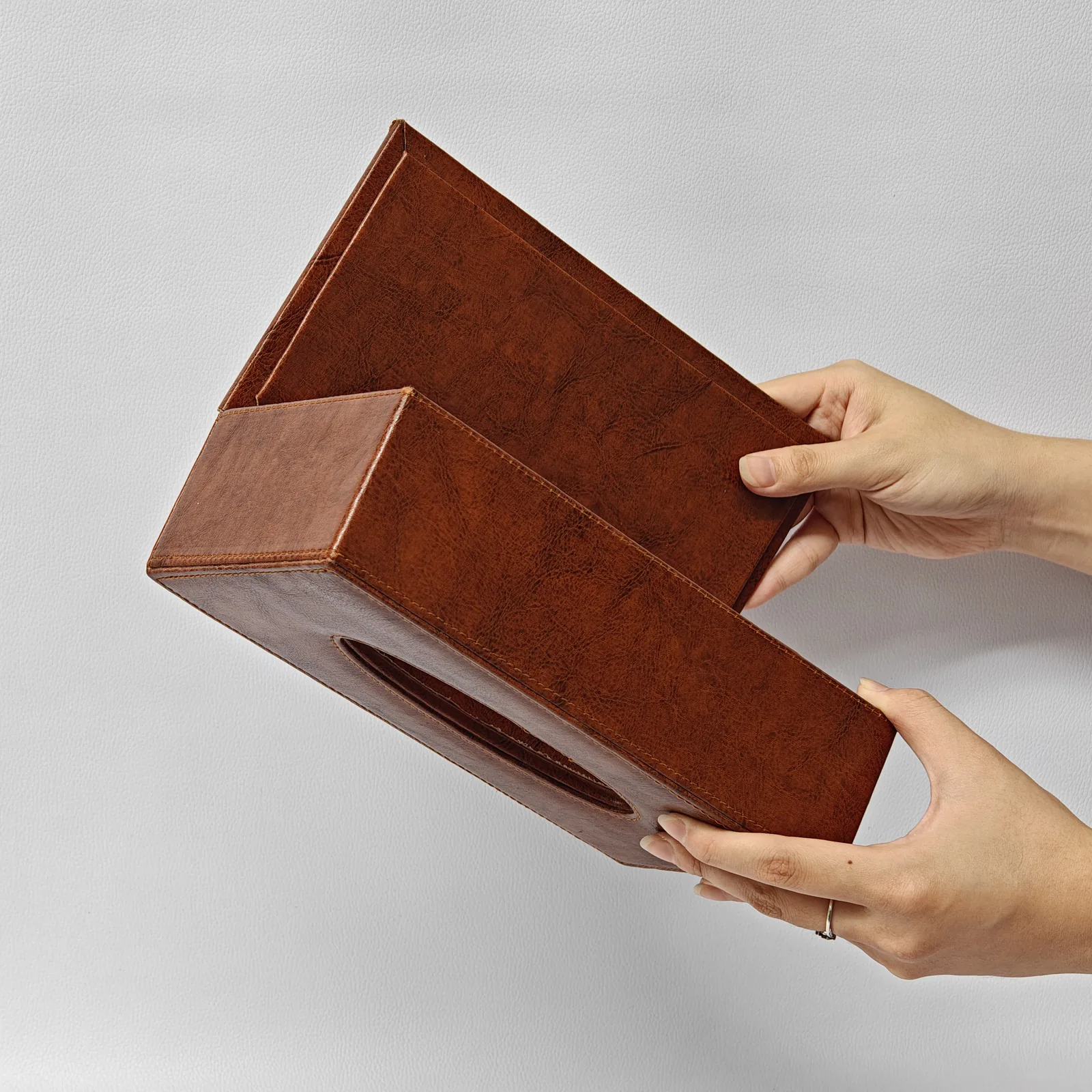 A hand holding a textured brown leather tissue box cover with the bottom flap fully open.