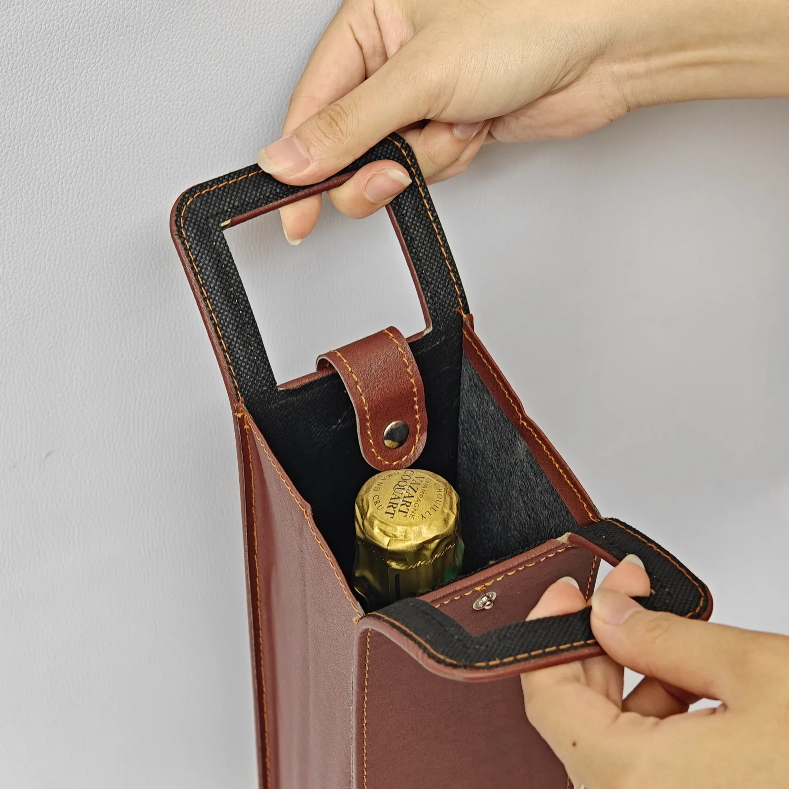 Hands holding open a brown leather wine carrier to show a champagne bottle inside.