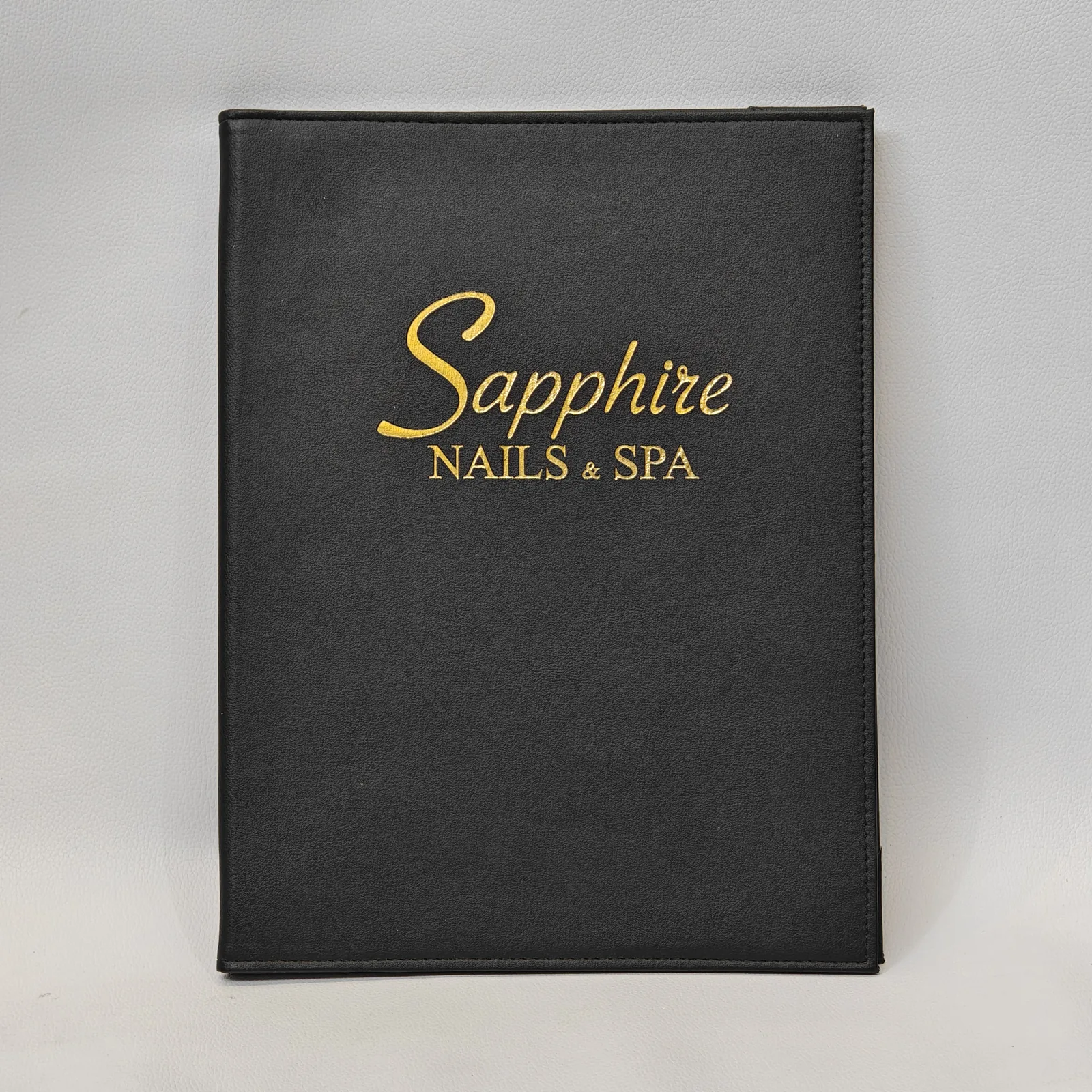 Front view of a black pebbled leather menu cover with gold foil text and perimeter stitching.