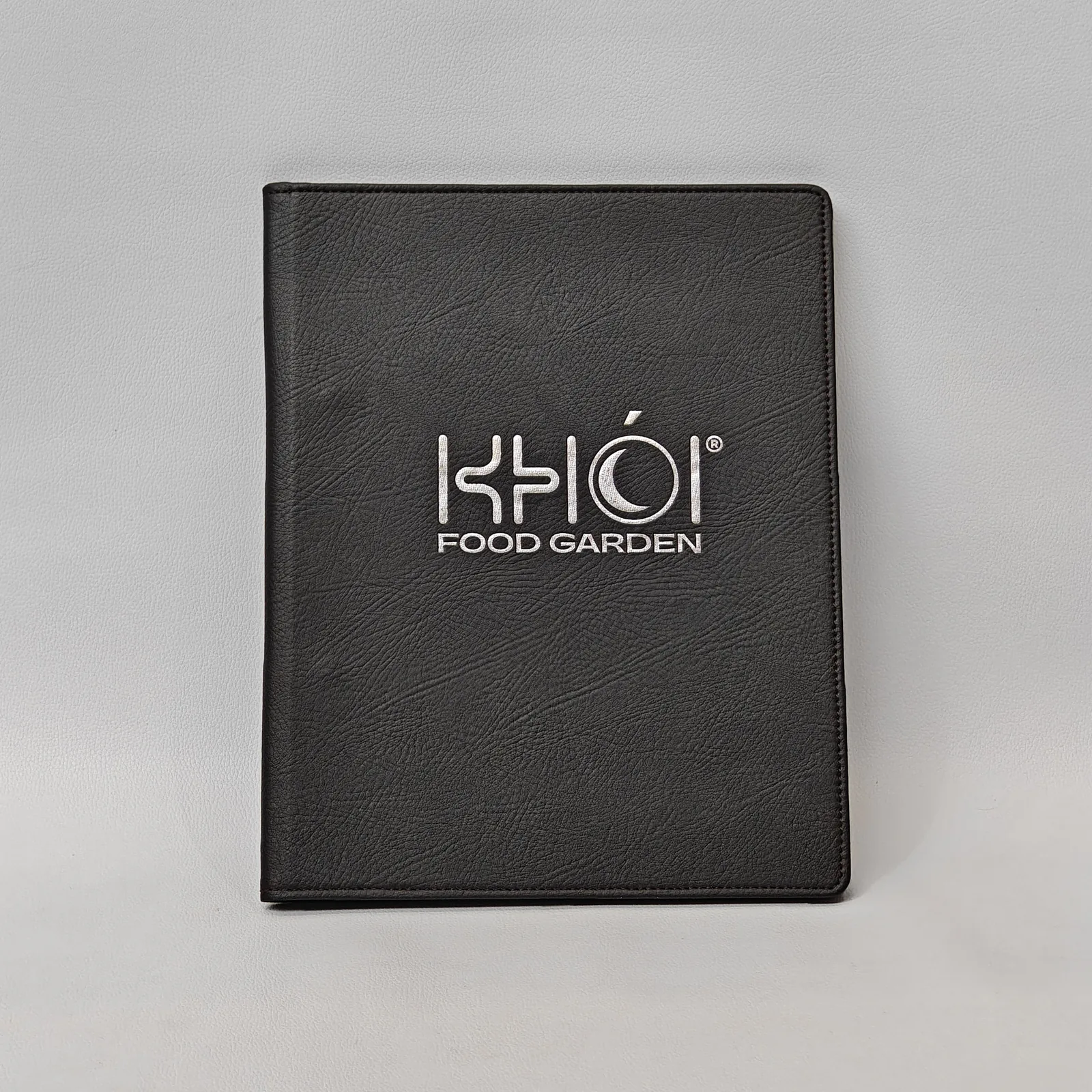 Front view of a dark brown textured leather menu binder with a silver logo.