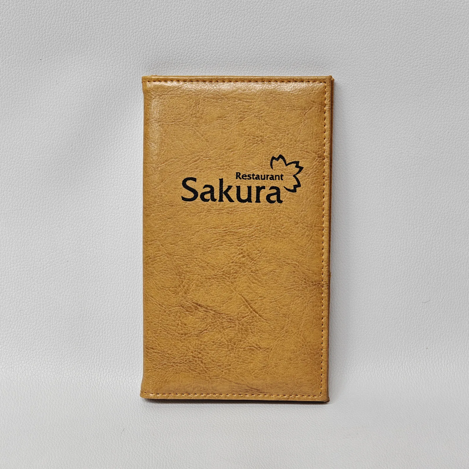 Front view of a closed tan leather check presenter with a black graphic logo and perimeter stitching.